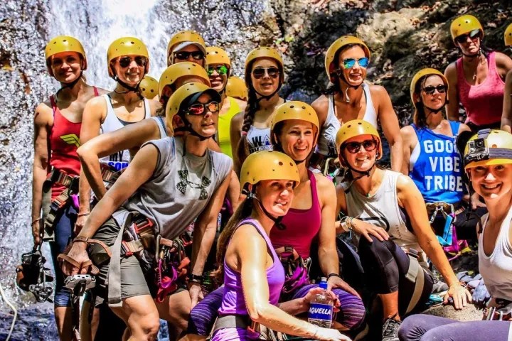 A group of people wearing helmets and climbing gear pose outdoors with a waterfall in the background.