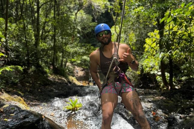 Person rappelling down a waterfall in a forest, wearing a helmet and smiling.