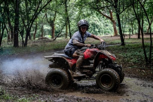 Person riding a red ATV through a muddy forest trail, splashing mud, wearing a helmet.