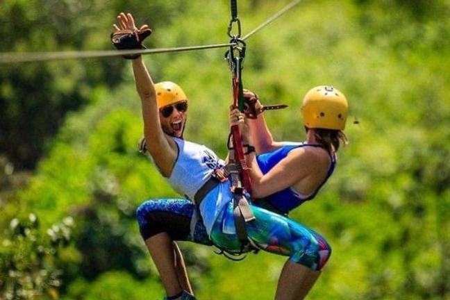 Two people ziplining together, wearing helmets and colorful outfits, one person waving excitedly.