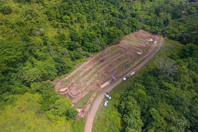 Aerial view of terraced farmland surrounded by dense green forest with a dirt road and parked vehicles.