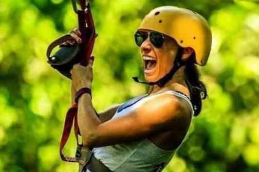 Person wearing helmet and sunglasses zip-lining through a forest, expressing excitement.