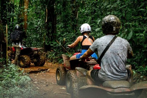 Three people riding ATVs on a narrow forest trail surrounded by green foliage.