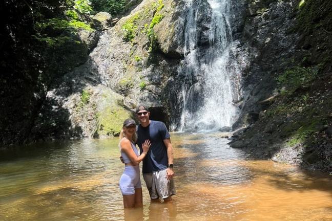 Couple standing in shallow water in front of a waterfall in a forest setting.