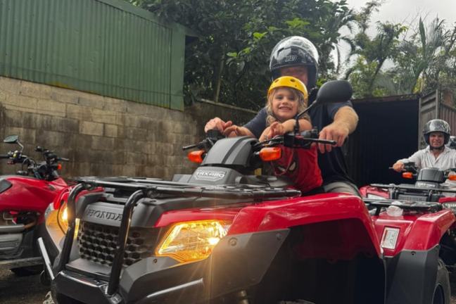 Child and adult riding a red ATV with helmets, surrounded by greenery and buildings.