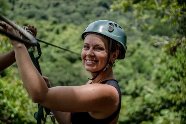 Smiling person in helmet ziplining with lush green forest background.