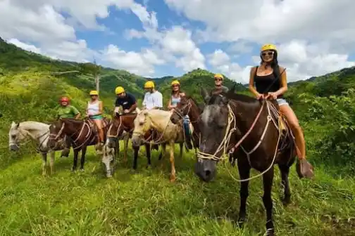 Group of people horseback riding on a grassy hillside with helmets under a blue sky.
