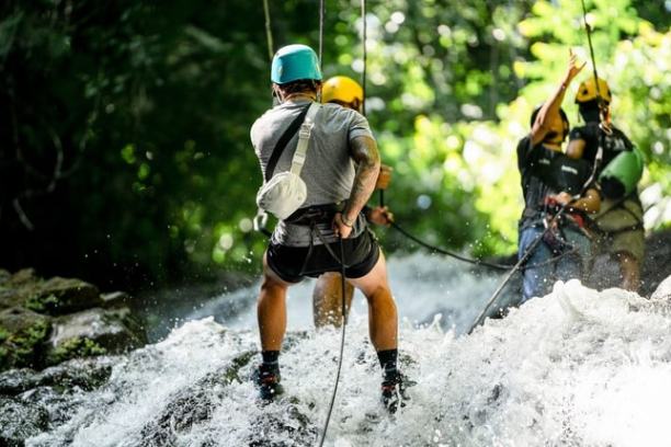Three people rappelling down a waterfall, wearing helmets and harnesses, surrounded by greenery.