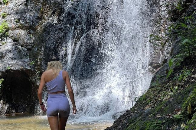 Person in shorts walking toward a waterfall in a lush, rocky setting.