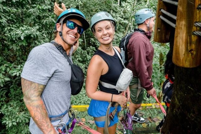 Two people in helmets and harnesses smiling, standing on a platform in a forest canopy.