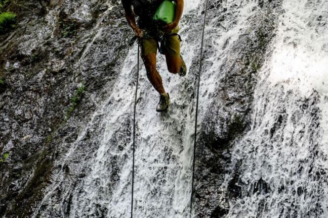 Person rappelling down a waterfall, wearing a yellow helmet and holding green equipment.
