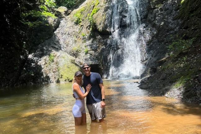 Couple standing in a shallow pond with a waterfall and lush greenery in the background.