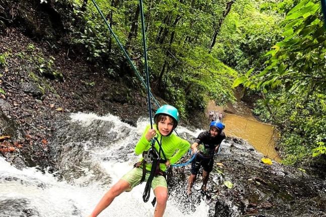 Two people rappelling down a waterfall with helmets and ropes in a lush forest.