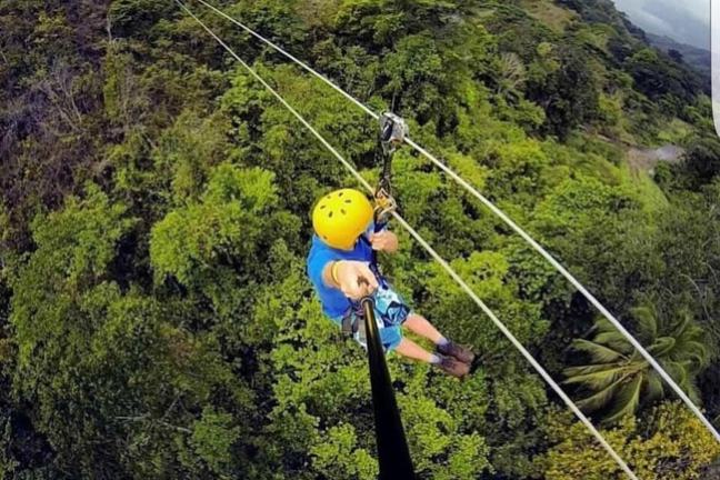 Person ziplining through lush forest, wearing a yellow helmet and holding a camera stick.