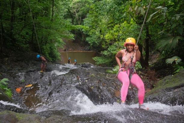 Person in pink pants rappels down a waterfall in a lush forest, wearing a yellow helmet.