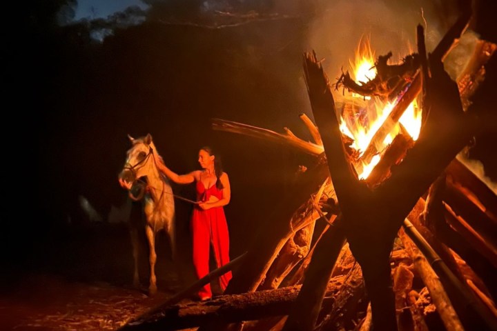 Person in red dress with horse near large bonfire at night, surrounded by trees.