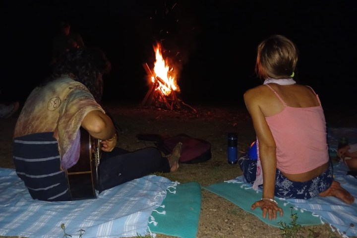 Two people sitting on blankets near a campfire at night, one playing guitar.
