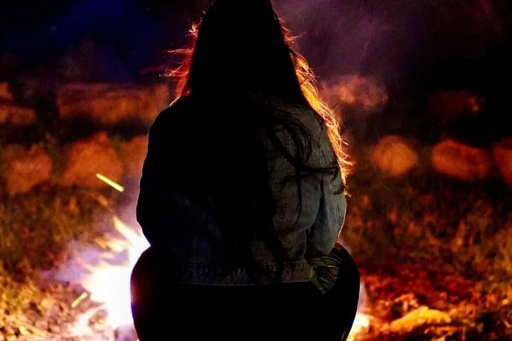 Silhouette of a person squatting by a campfire at night.