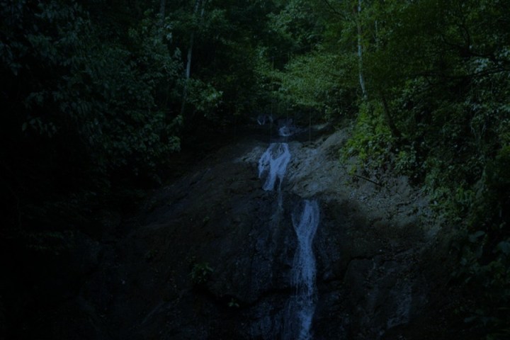 Dimly lit waterfall surrounded by dense green foliage and rocky terrain.